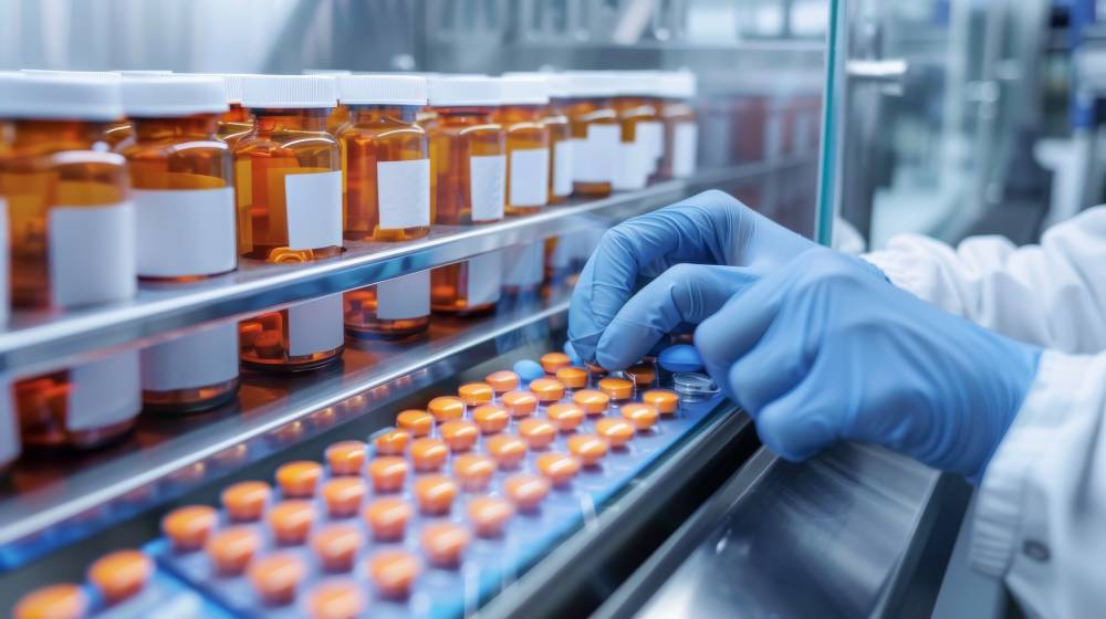 A gloved hand handles orange pills on a production line beside rows of amber pill bottles with white labels, conveying supply chain and manufacturing of pharmaceuticals.