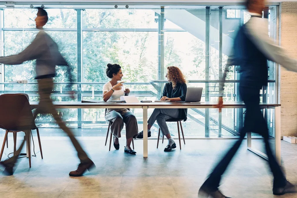 Two women colleagues collaborating in a busy office, conveying a collaborating work environment.